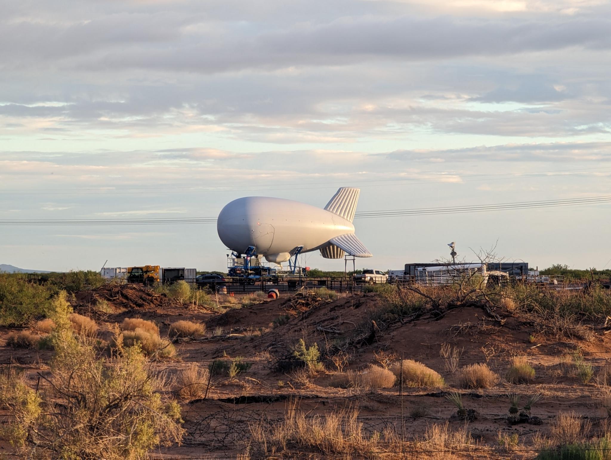 The Tactical Aerostat System at Santa Teresa Station. Image Courtesy of Battalion Search and Rescue A white blimp on the ground in the middle of the desert.